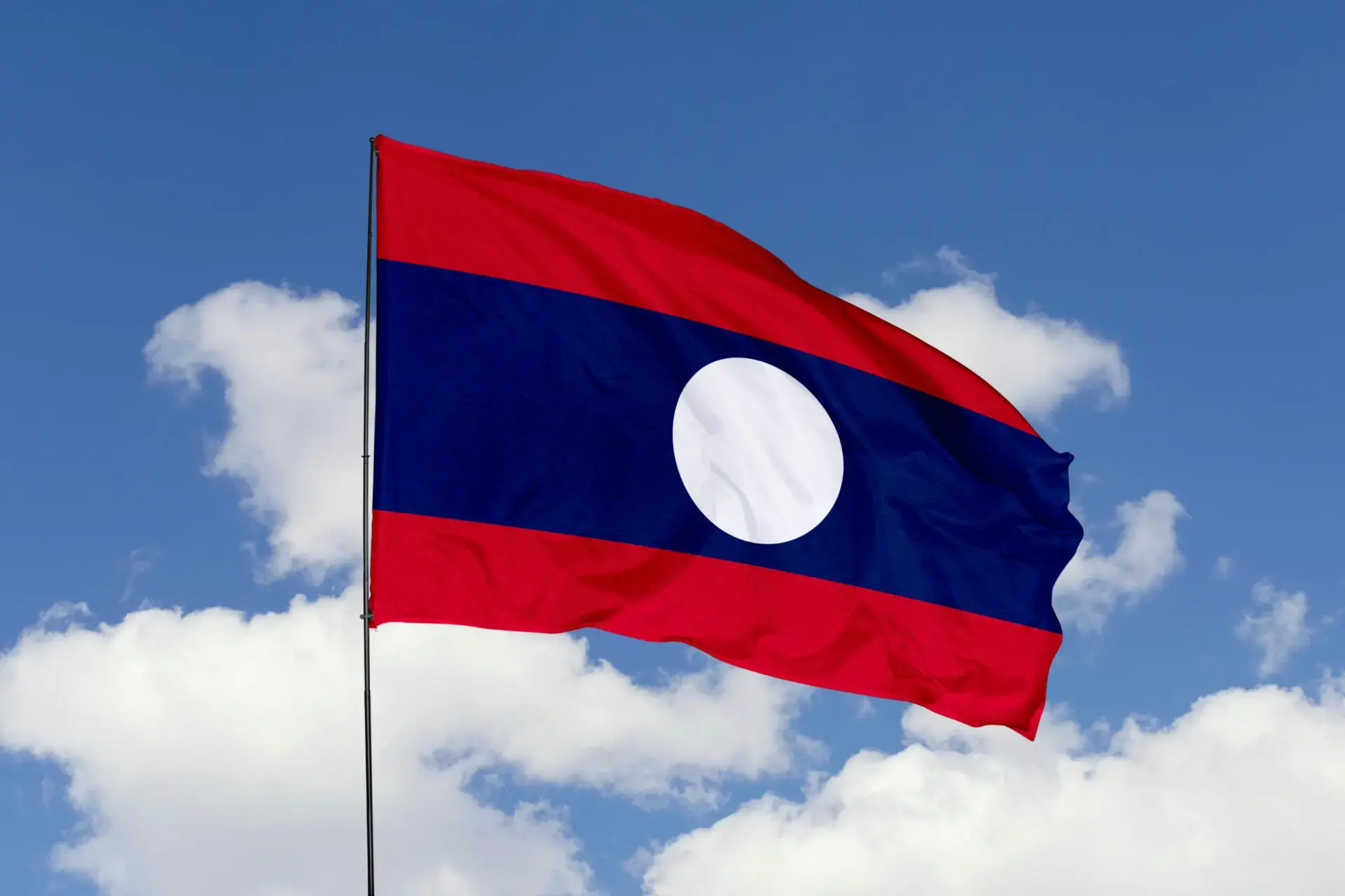 The flag of Laos, featuring three horizontal stripes—red, blue, and red—with a large white circle centered on the blue stripe, waves against a blue sky with scattered clouds.