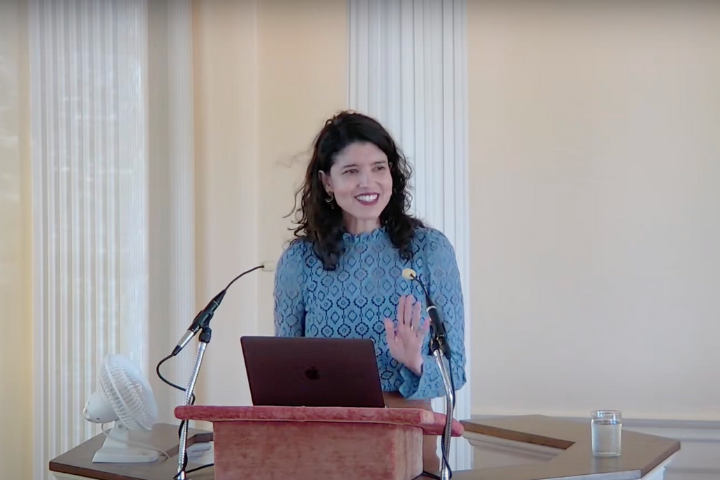 A woman with dark hair, wearing a blue patterned dress, stands at a podium with a laptop and microphones, smiling and gesturing with her hand in a bright room with white columns.