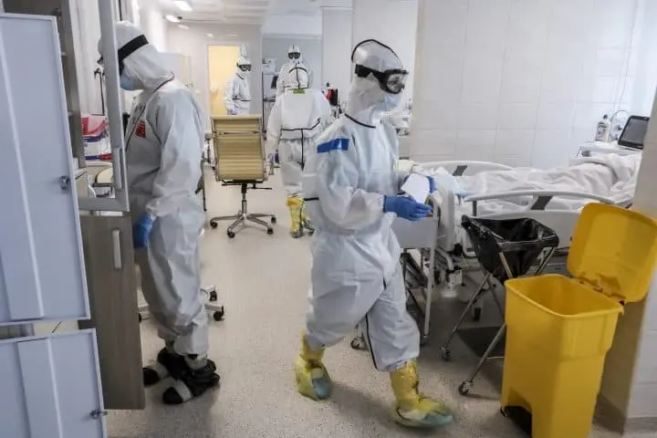 Medical workers in Eurasia wearing full protective suits, masks, and gloves care for COVID-19 patients in a hospital room. One patient lies in bed as staff attend to tasks, with medical equipment and a yellow waste bin visible.