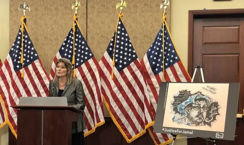 A woman speaks at a podium in front of several American flags; beside her is a display with artwork and the hashtag #JusticeforJamal, highlighting calls for accountability after Jamal Khashoggi’s death.