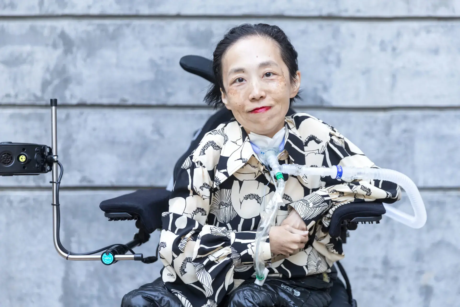 A woman with short hair sits in a motorized wheelchair, wearing a patterned shirt and dark pants. She has a tracheostomy tube and is smiling in front of a gray stone wall.