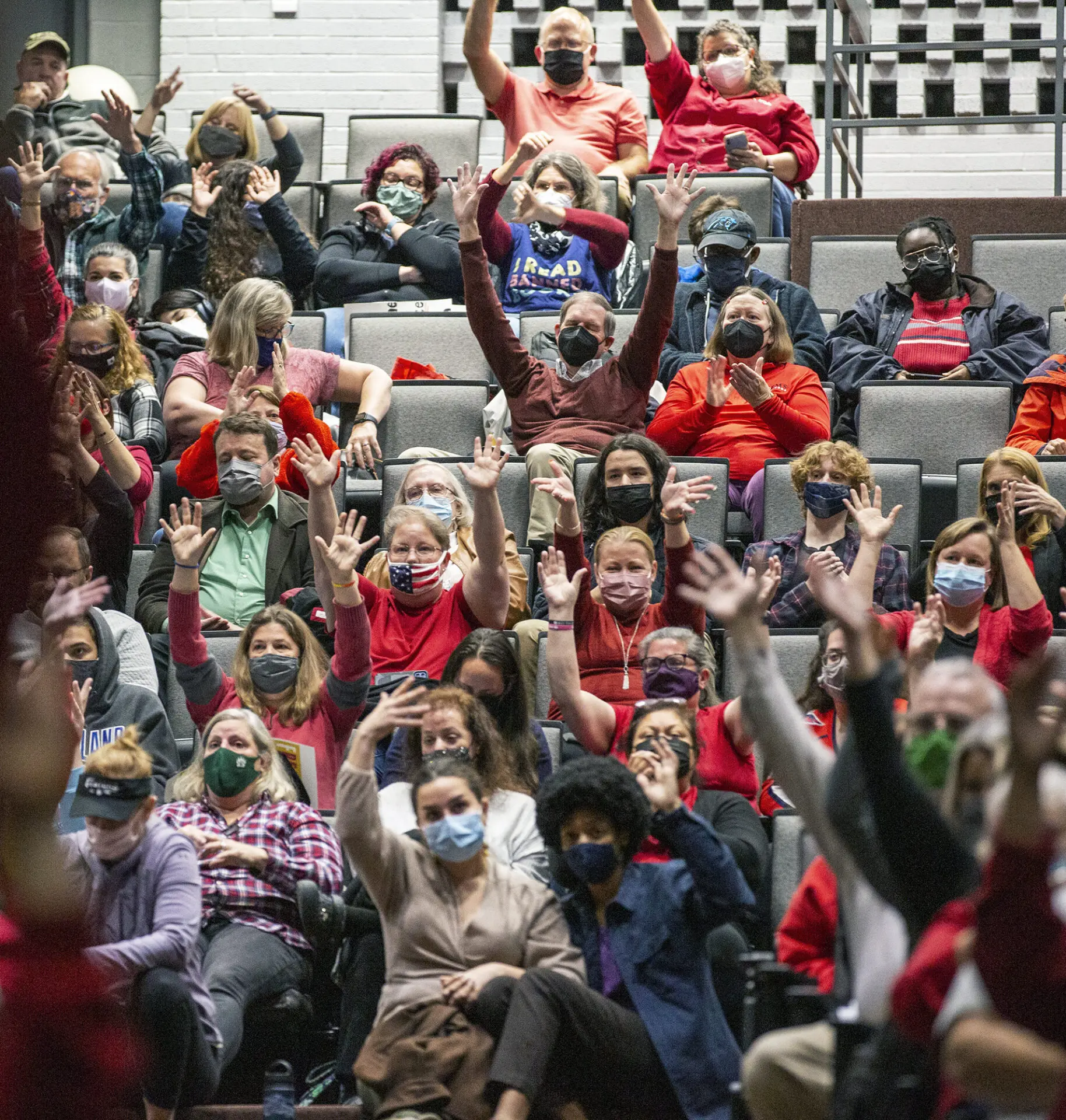 A large group of people wearing masks and casual clothes sits in theater seats, many raising their hands enthusiastically. The crowd includes diverse ages and ethnicities, with most people wearing red or brightly colored clothing, supporting Book Ban Resources.