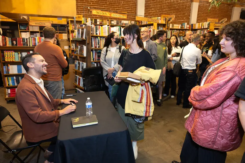 A smiling author sits at a table signing books for fans in a bookstore. People stand in line holding books, waiting to meet him, surrounded by shelves full of books.