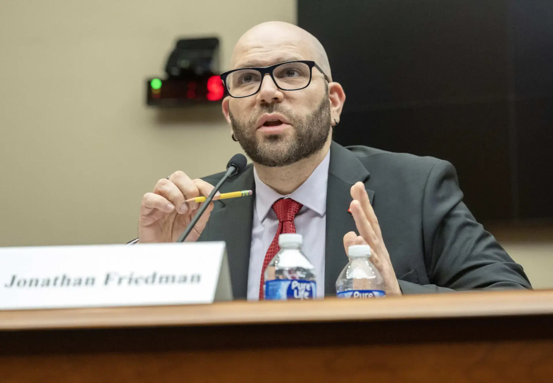 A man in a suit and red tie speaks into a microphone at a hearing, holding a pencil. A nameplate reading Jonathan Friedman and two water bottles are in front of him.