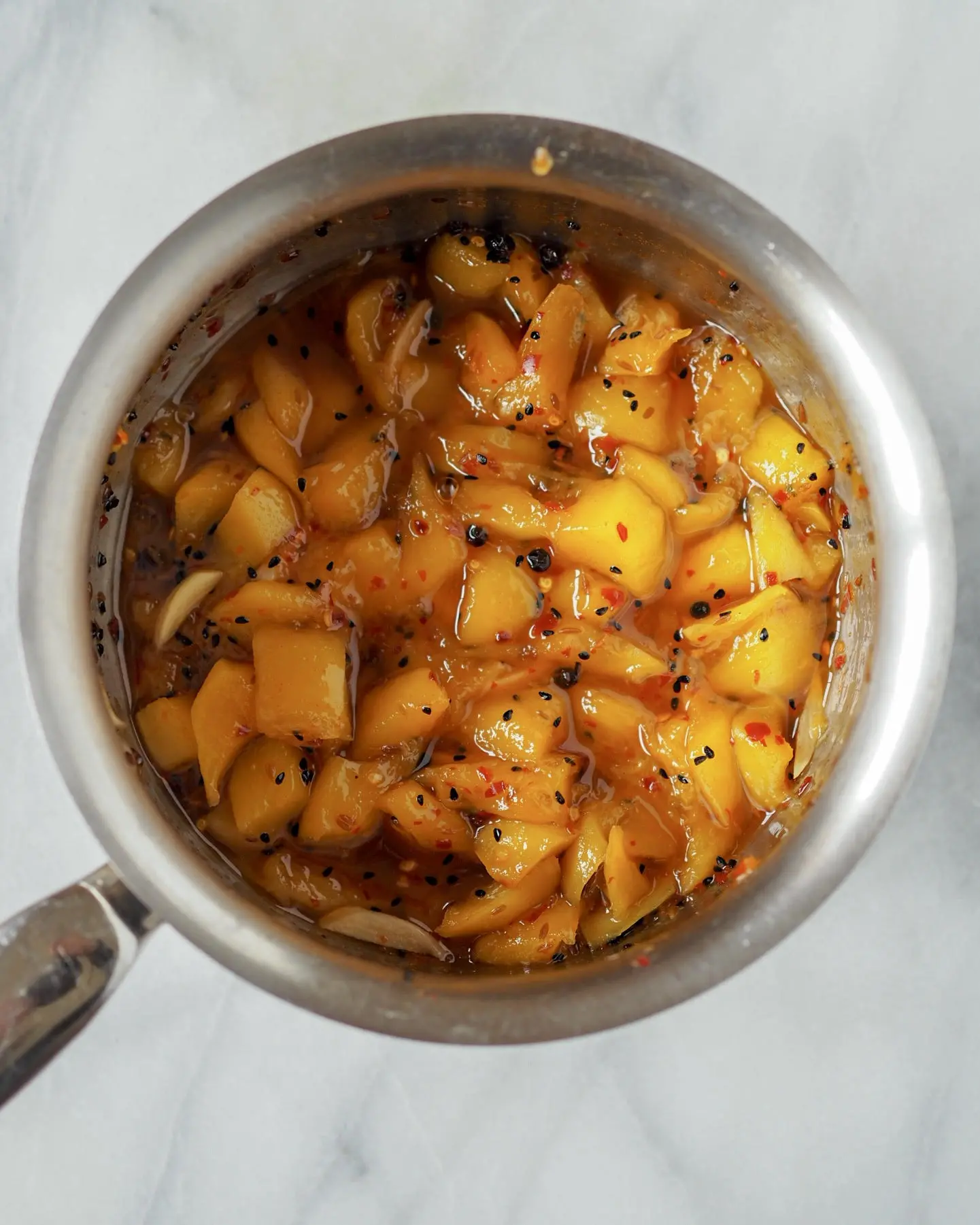A saucepan filled with chunky mango chutney, inspired by Pooja Makhijani cooking, features pieces of mango, seeds, and spices in a glossy, syrupy mixture, viewed from above on a light marble surface.