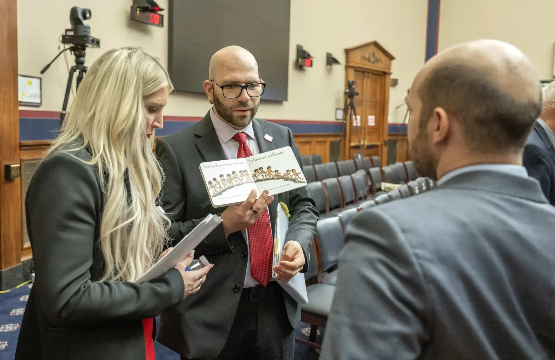 A woman and two men in business attire talk in a conference room. One man holds a paper with photos and text. Camera equipment and empty chairs are visible in the background.