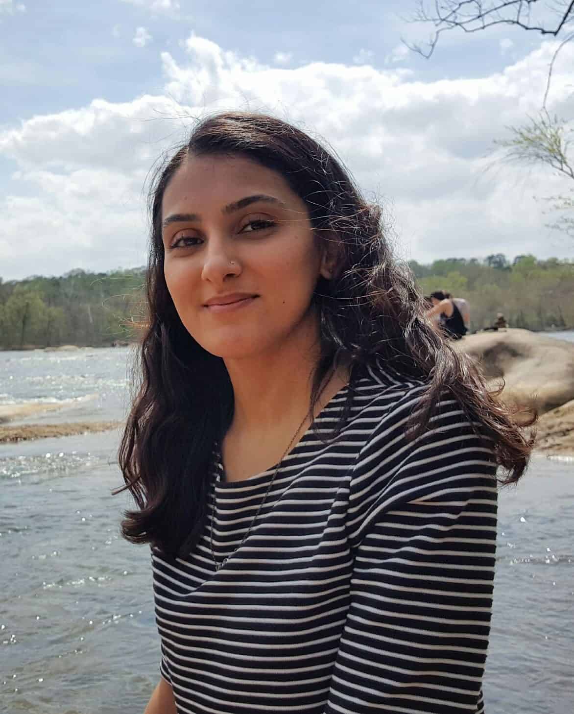 A Muslim woman in America with long dark hair, wearing a black and white striped shirt, sits by a river with trees and a cloudy sky in the background. She smiles gently at the camera.