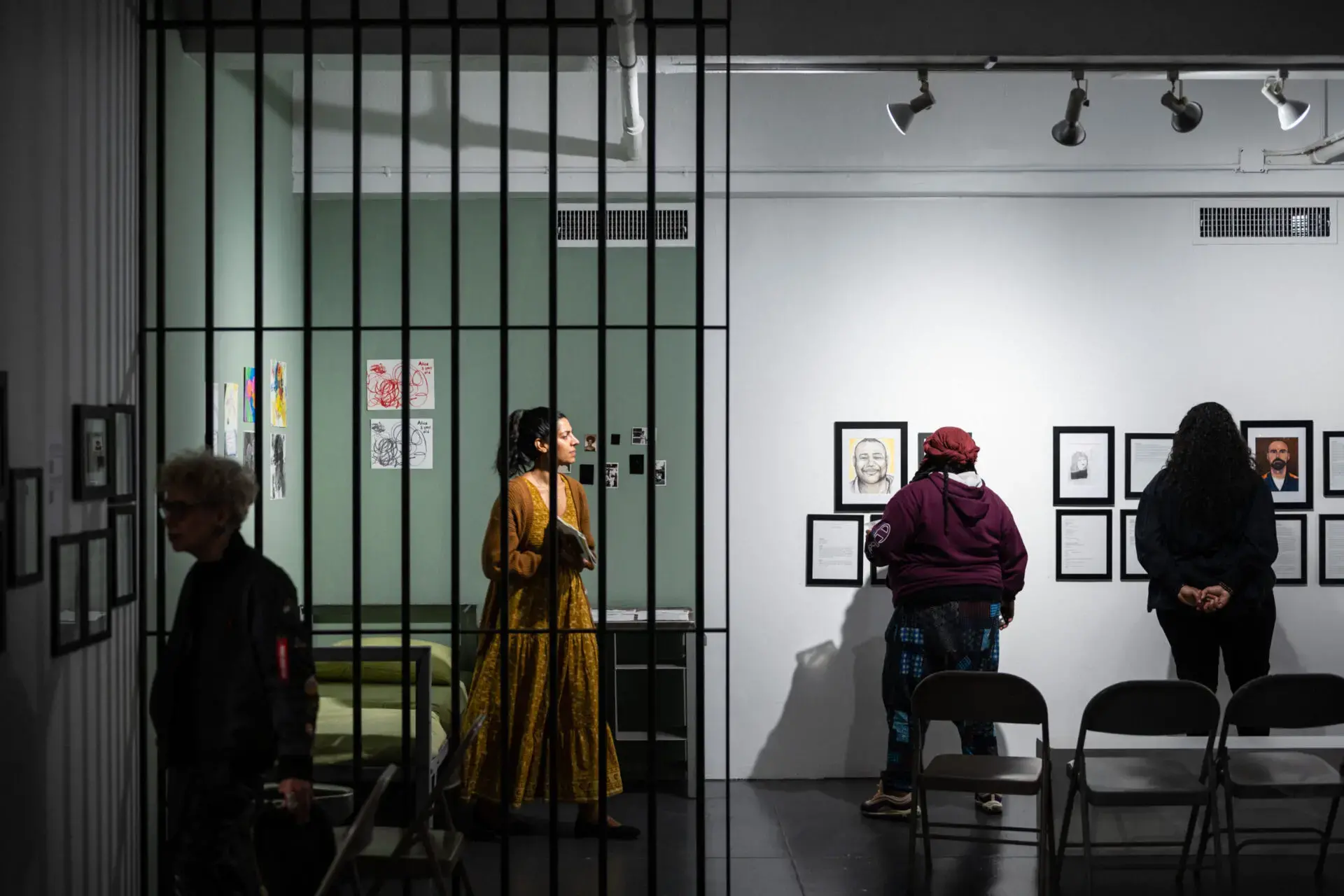 Several people view artwork on the walls of a gallery, seen through metal bars; one woman in a yellow dress stands in the center, while others face framed pieces under bright lights.
