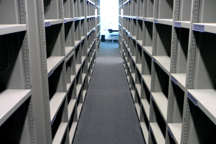 Rows of empty gray metal shelves line both sides of a narrow aisle in a well-lit room, with a desk and chair visible at the far end near a window.