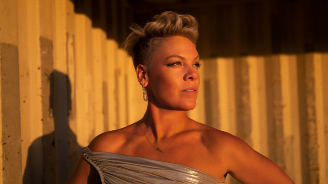 A person with short, styled blonde hair stands in sunlight, wearing a one-shoulder metallic dress and earrings, gazing confidently into the distance against a backdrop of vertical panels.