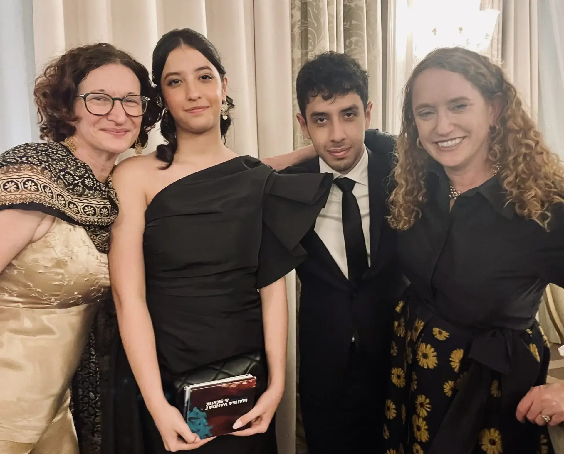Four people dressed in formal attire stand together, posing and smiling for a photo at an indoor event. Two women on the left and right, and a woman and man in the center, all appear happy and well-dressed.