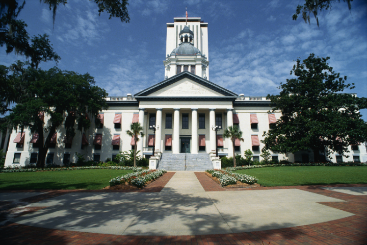 The image shows the front view of the Florida State Capitol building in Tallahassee, with a central dome, white columns, red-striped awnings, and landscaped gardens under a blue sky with scattered clouds.