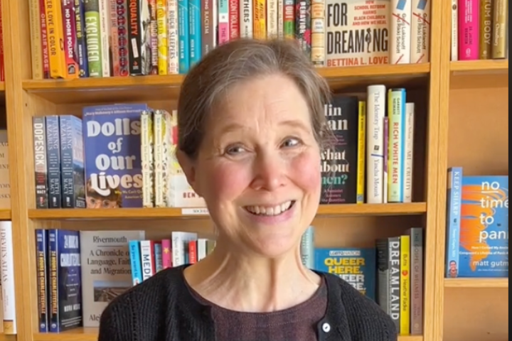 A woman with short brown hair and a warm smile stands in front of bookshelves filled with colorful books. She is wearing a dark top and a black cardigan.
