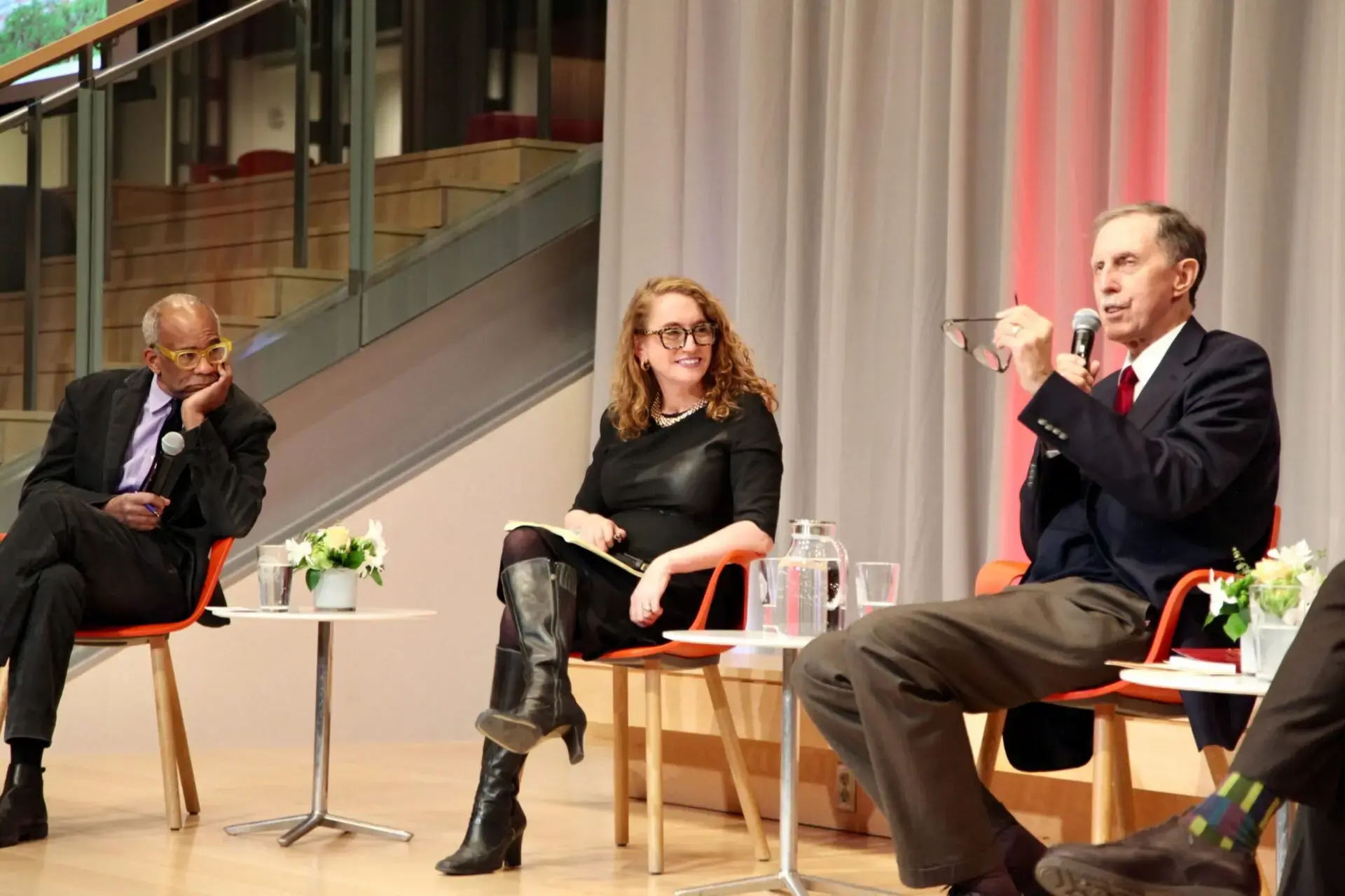 Three people sit on stage in chairs during a panel discussion. One man gestures while speaking, a woman in black dress and boots smiles, and another man listens with his hand on his face. Small tables with flowers are beside them.
