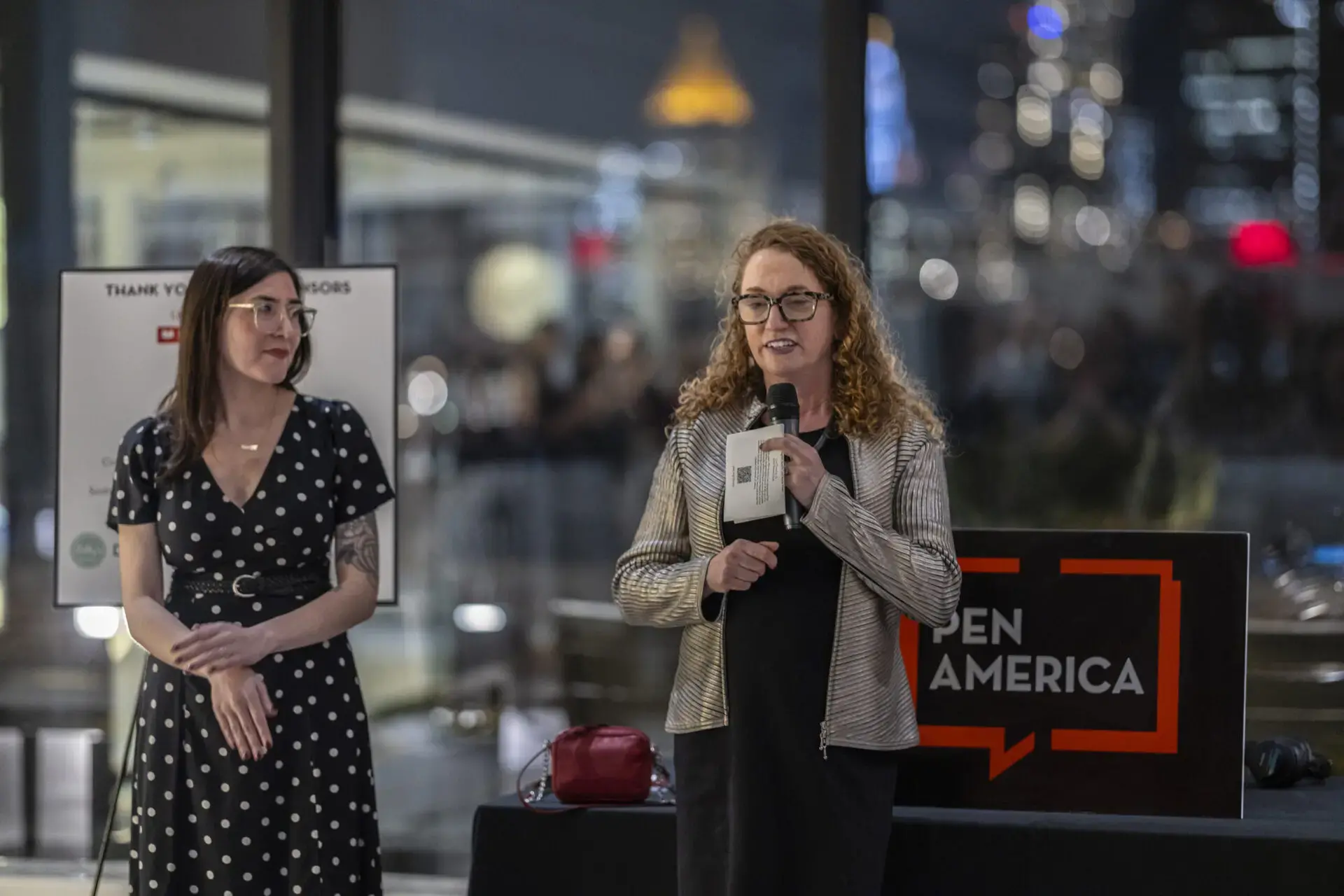 Two women stand indoors at an event; one speaks into a microphone while holding papers, the other stands nearby. A sign reads PEN America and city lights are visible through large windows behind them.