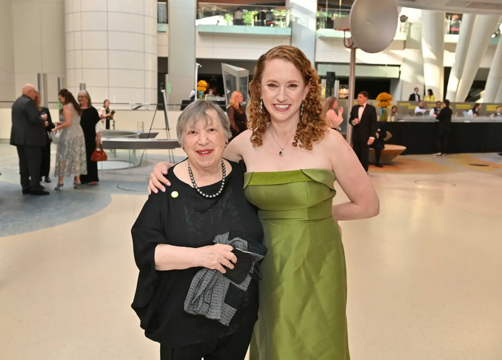 Two women pose together at an indoor event; one wears a black outfit with a beaded necklace, and the other wears a green strapless dress. People and tables are visible in the background.