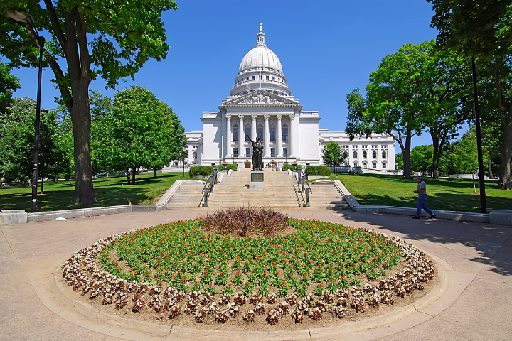 A large white capitol building with a prominent dome stands behind a circular flower bed, trees, and a walkway under a clear blue sky. A few people are walking on the grounds.