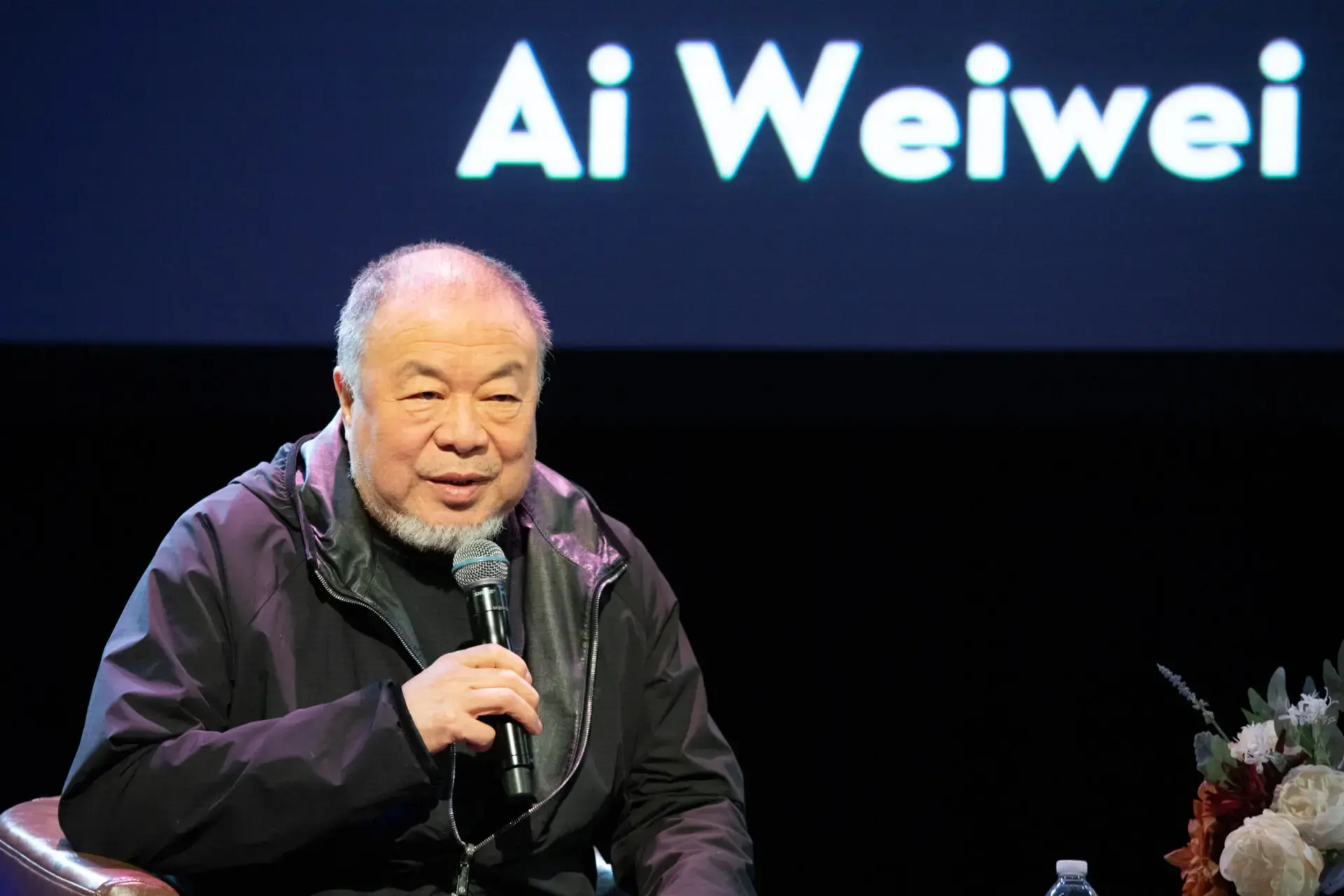 An older man with a beard speaks into a microphone while seated onstage. Behind him, the name Ai Weiwei is displayed in large white letters on a dark background.