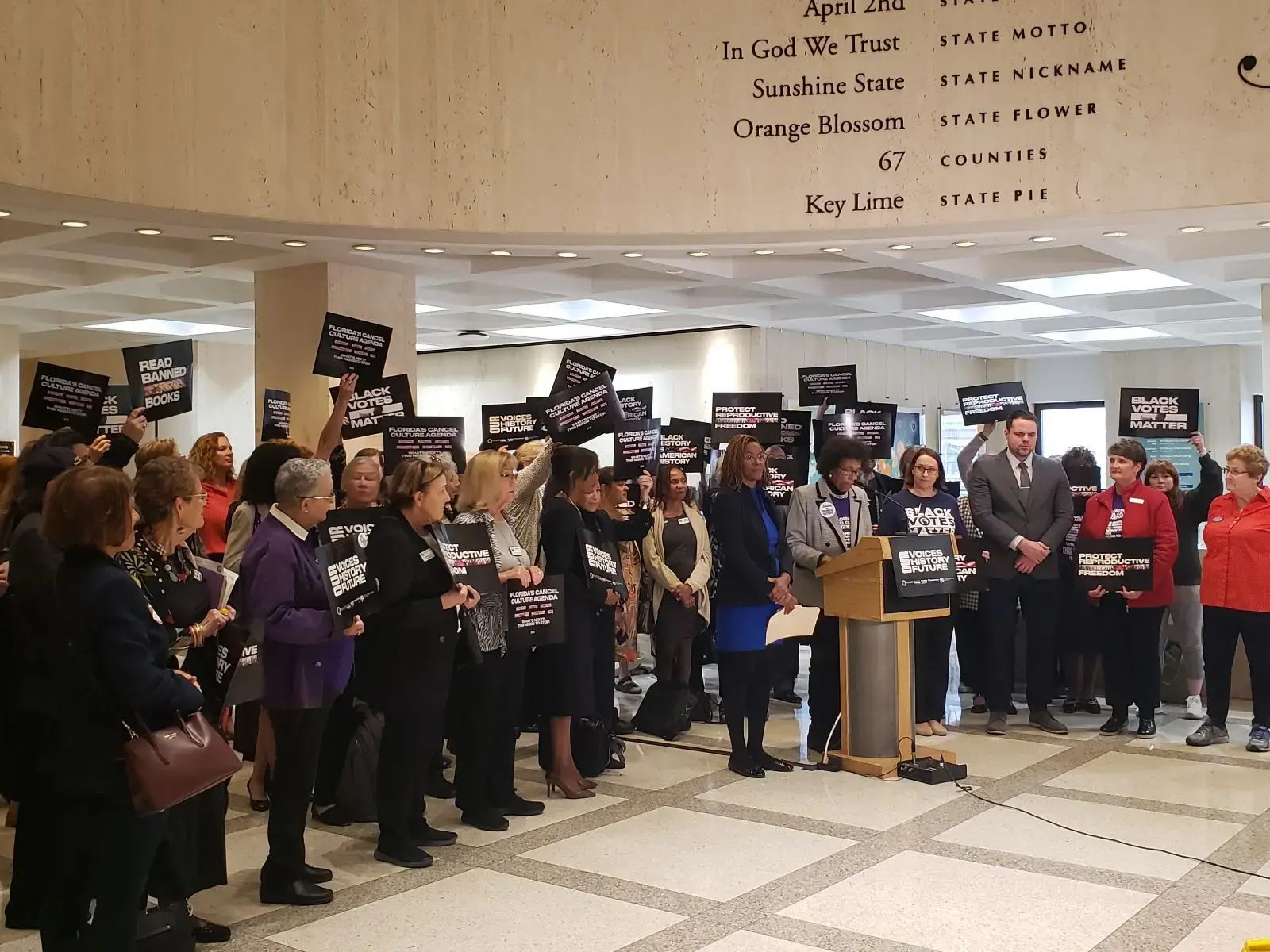 A diverse group of people stand indoors, many holding signs that read “BLACK HISTORY IS AMERICAN HISTORY.” A woman speaks at a podium, while others listen. Text about Florida is visible on the wall above.