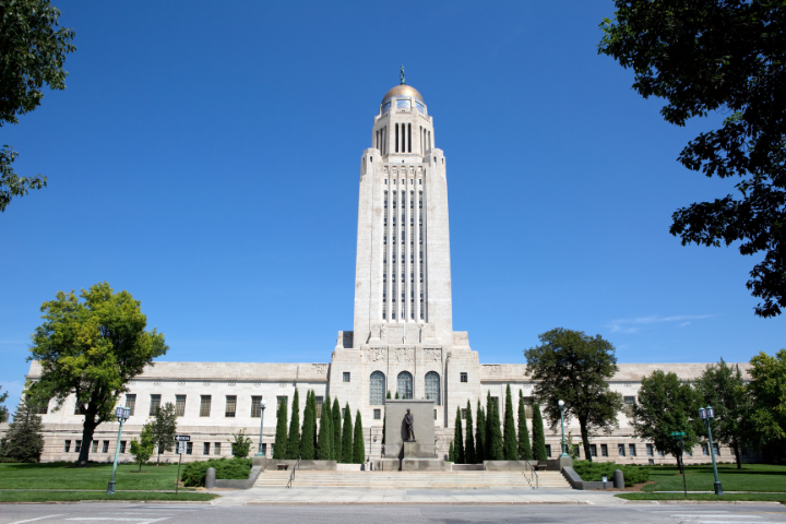 The Nebraska State Capitol building in Lincoln, featuring a tall central tower with a golden dome, surrounded by trees and a clear blue sky.