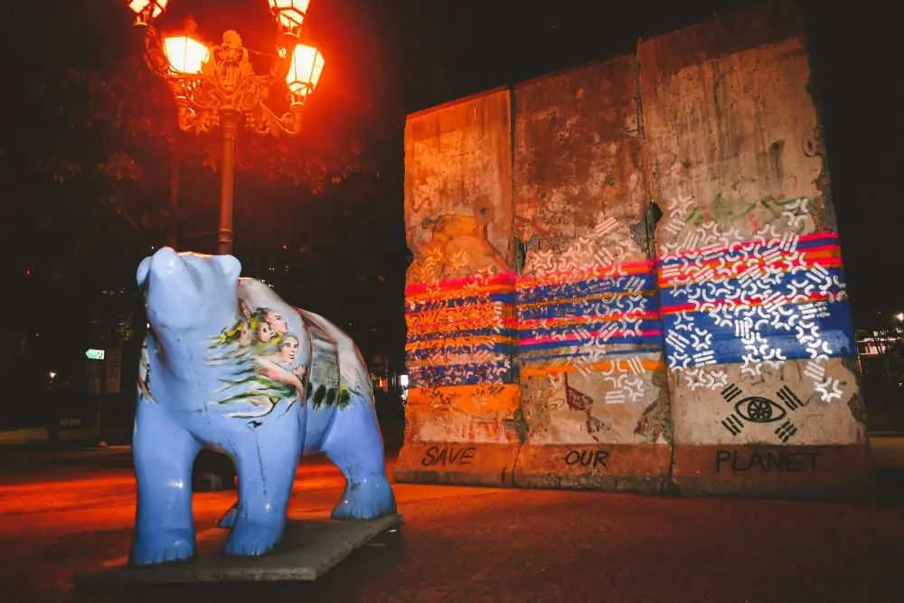 A painted bear sculpture and two Berlin Wall segments, featuring HIDEYES graffiti artist’s work, stand under streetlights at night. The words “Save Our Planet” and the South Korean flag are visible on the wall.