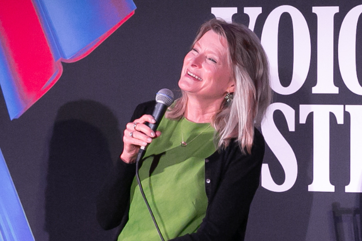 A woman with blonde hair, wearing a green top and black cardigan, smiles and holds a microphone while performing on stage in front of a partially visible sign.