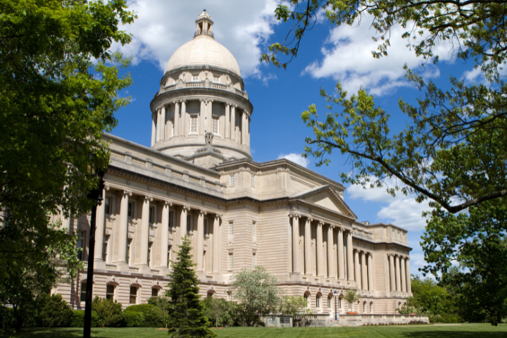 A large, classical-style government building with tall columns and a central dome, surrounded by green trees and grass under a blue sky with scattered clouds.