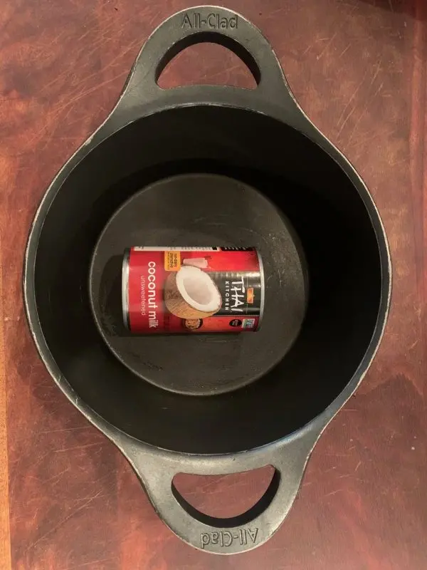 A can of coconut milk sits inside a large, empty black All-Clad pot on a wooden surface, ready for a James Sie cooking session.