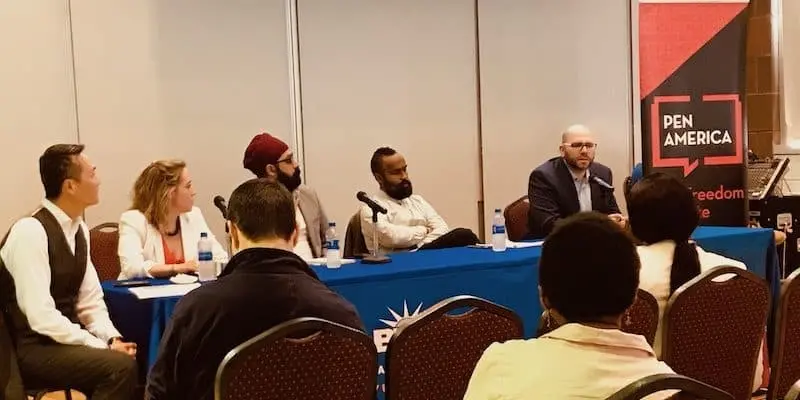A panel of five people sits at a blue table with microphones, engaging in a campus free speech conversation with the audience. A red and black PEN America banner is visible in the background.