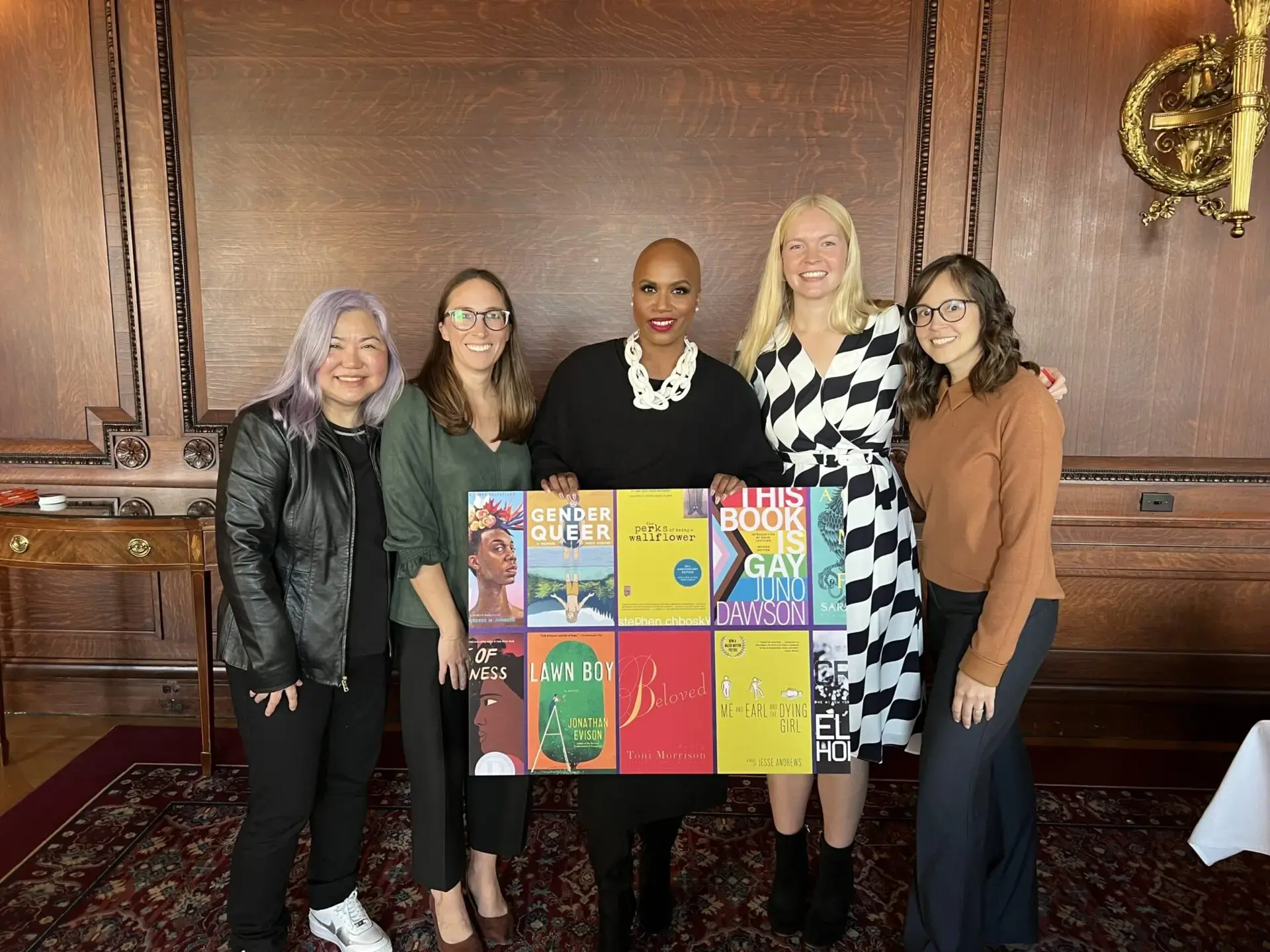 Five people stand together, smiling, in a wood-paneled room. The person in the center holds a display of colorful book covers, including titles like Gender Queer and This Book Is Gay.