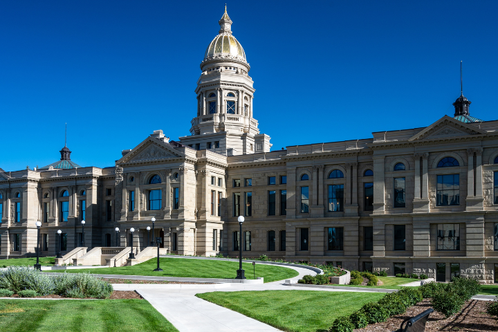 The photo shows a grand stone building with a golden dome, arched windows, and columns, set against a clear blue sky. Manicured lawns and walkways lead up to the entrance.