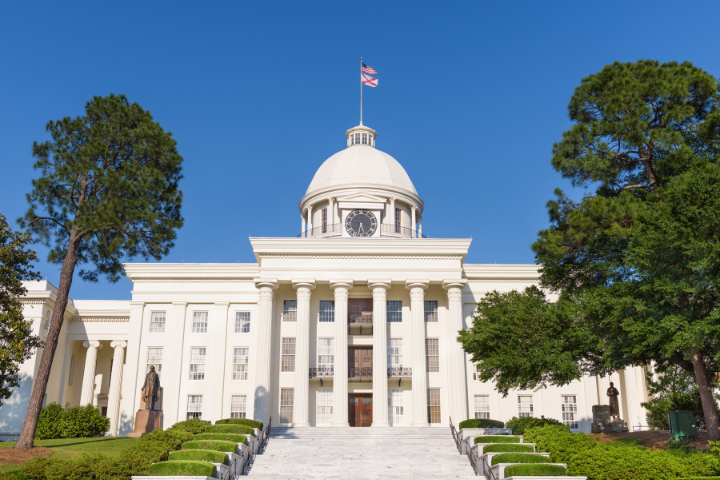 The Alabama State Capitol building in Montgomery, featuring a white dome, columns, clock, and a U.S. flag on top, surrounded by trees under a clear blue sky.