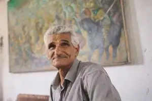 An elderly man with gray hair and a mustache, wearing a gray shirt, sits indoors with a gentle expression. A colorful, blurred painting hangs on the wall behind him.