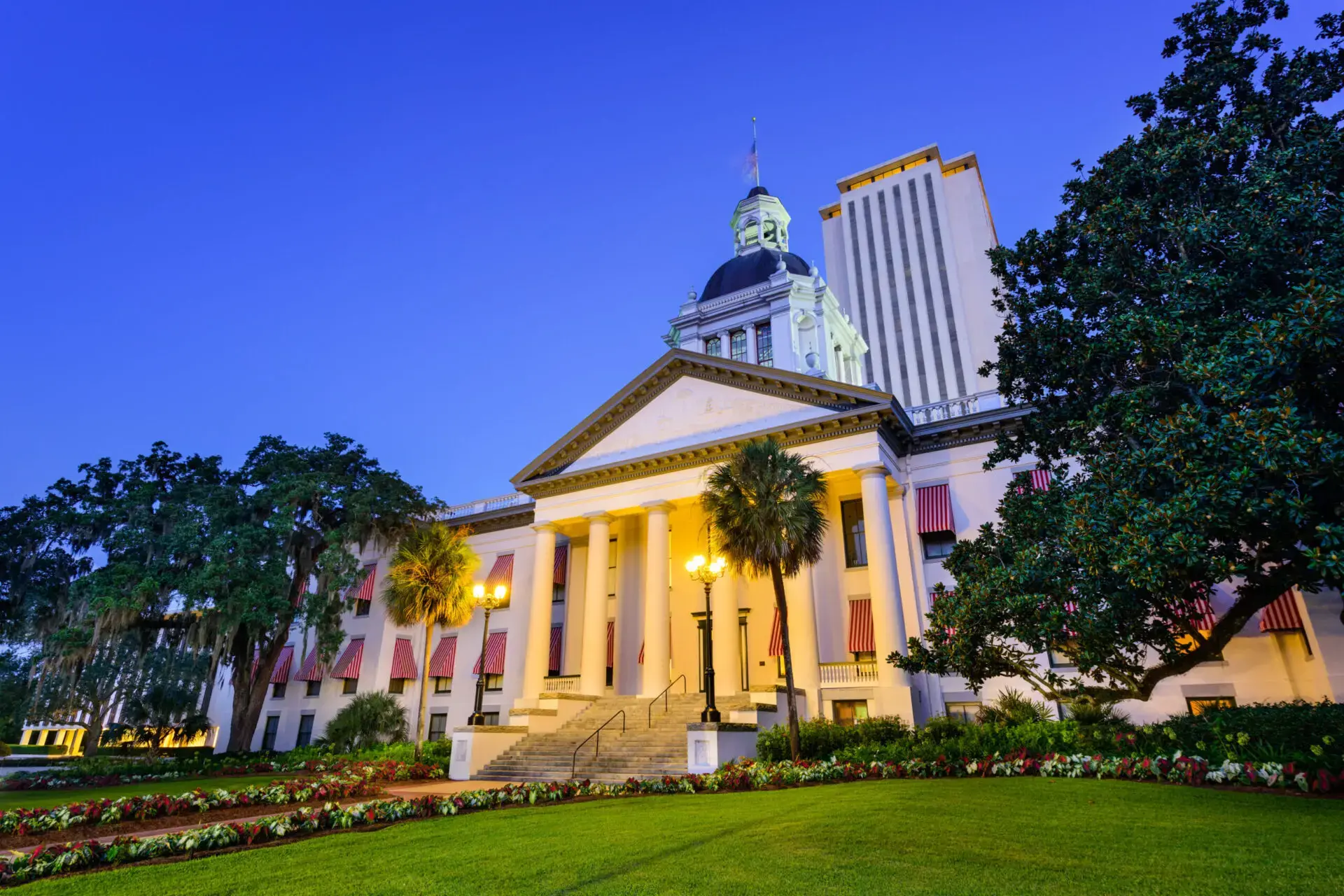 White government building with tall columns and a dome, lit up at dusk, surrounded by palm trees, green lawn, colorful flowers, and modern skyscraper in the background.