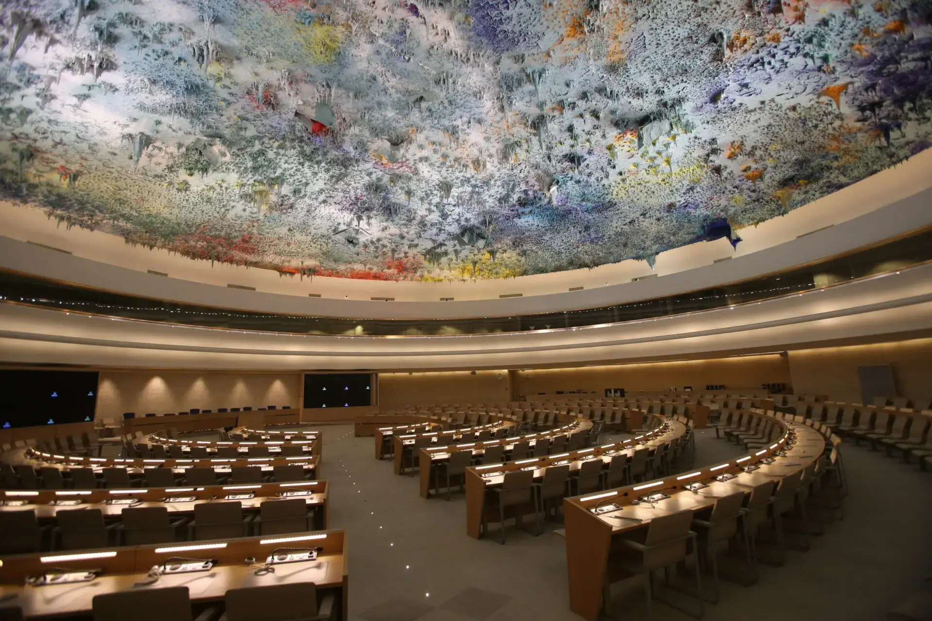 A large, empty conference hall with curved rows of desks and chairs, microphones on each desk, and a colorful, abstract painted ceiling. The room is well-lit and modern in design.