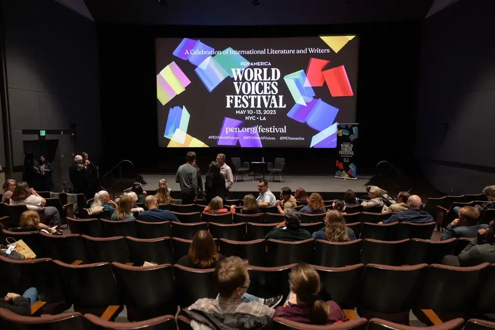 Audience members sit in a theater facing a large screen displaying colorful graphics and text for the PEN America World Voices Festival, held May 10-13, 2023, in NYC and LA. Some people are talking before the event starts.