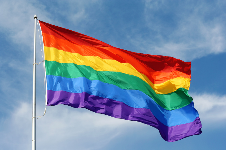 A rainbow pride flag with six horizontal stripes—red, orange, yellow, green, blue, and purple—waves on a flagpole against a blue sky with scattered white clouds.
