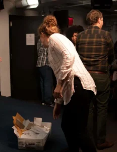 A woman in a white striped shirt bends over near a white bin filled with envelopes and papers, possibly related to writing in prison, while others stand and talk in a dimly lit room.