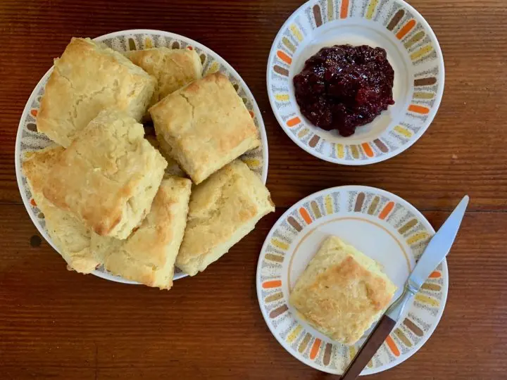 A plate of square biscuits sits on a wooden table next to a small dish of berry jam—an inviting scene inspired by Steven Reigns cooking—with a plate holding a single biscuit and butter knife nearby.