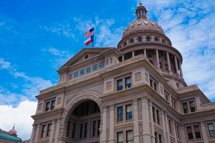The image shows the Texas State Capitol building in Austin, featuring its ornate dome and facade under a blue sky. The American and Texas flags are flying atop the structure.
