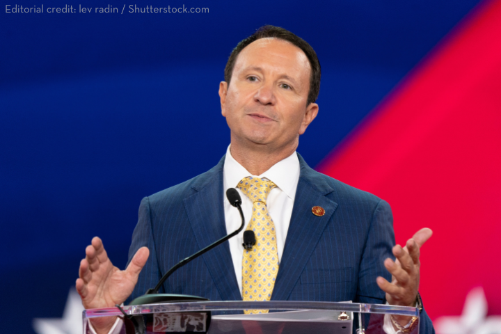 A man in a blue pinstripe suit and yellow tie speaks at a podium with microphones, gesturing with his hands. The background is red and blue with a subtle star design.