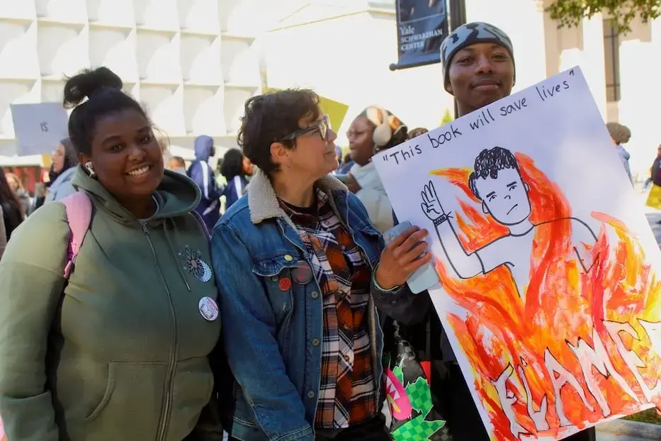 Three people stand together at a protest. One holds a sign with a drawing of a person in flames and the text This book will save lives. They are smiling, and others are visible in the background.