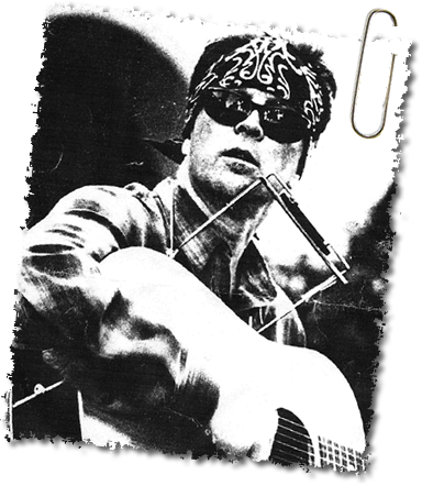 Black-and-white photo of a musician wearing sunglasses and a patterned bandana, playing an acoustic guitar and harmonica. The image has rough edges, a paperclip in the upper right corner, evoking the raw emotion of reentry into old rhythms.