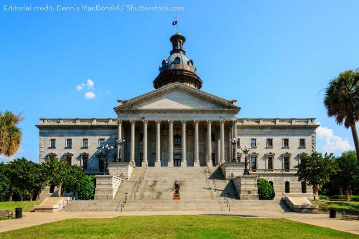 The South Carolina State House in Columbia, a grand neoclassical building with a large dome, columns, and a broad staircase, set against a clear blue sky and surrounded by green trees.