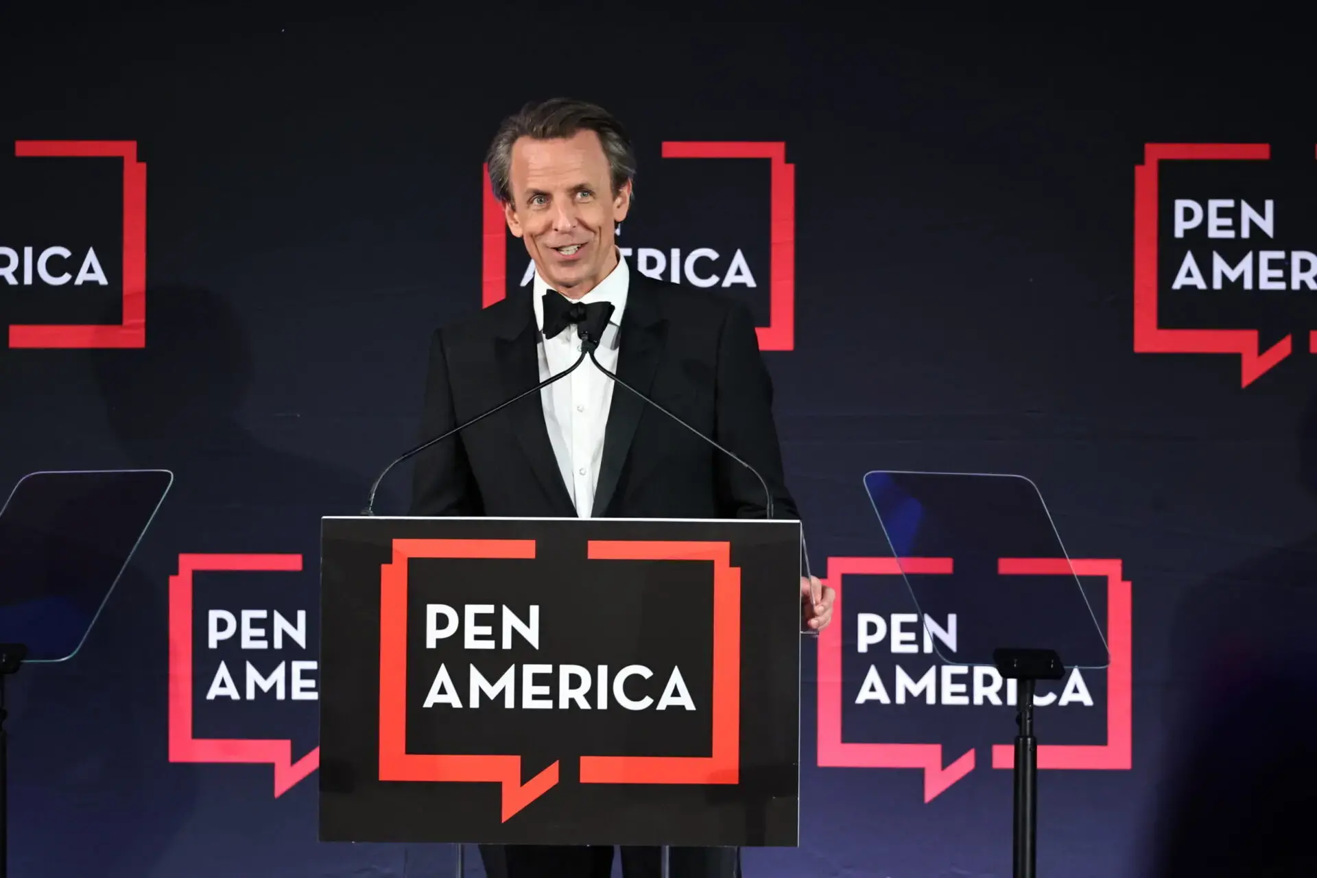 A man in a tuxedo stands at a podium with a microphone, speaking at a PEN America event. The podium and background display the PEN America logo with red speech bubbles.