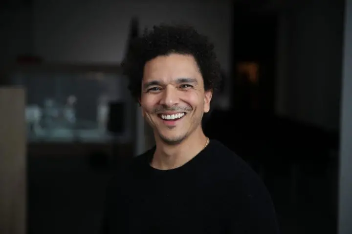 A man with curly hair and a goatee, wearing a black shirt, smiles warmly while standing indoors in a dimly lit space, evoking the inviting atmosphere found in "Unwrapping" by Damien Belliveau.