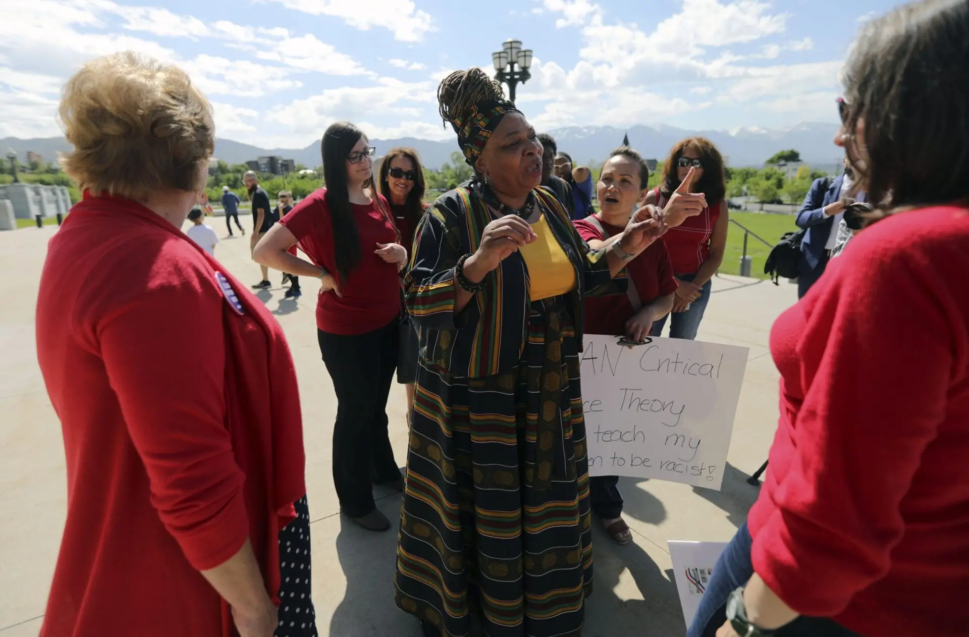 A woman in a patterned dress speaks passionately to a group, some in red shirts, outdoors on a sunny day. One person holds a sign about educational gag orders and the ban on critical race theory. Mountains and greenery are in the background.
