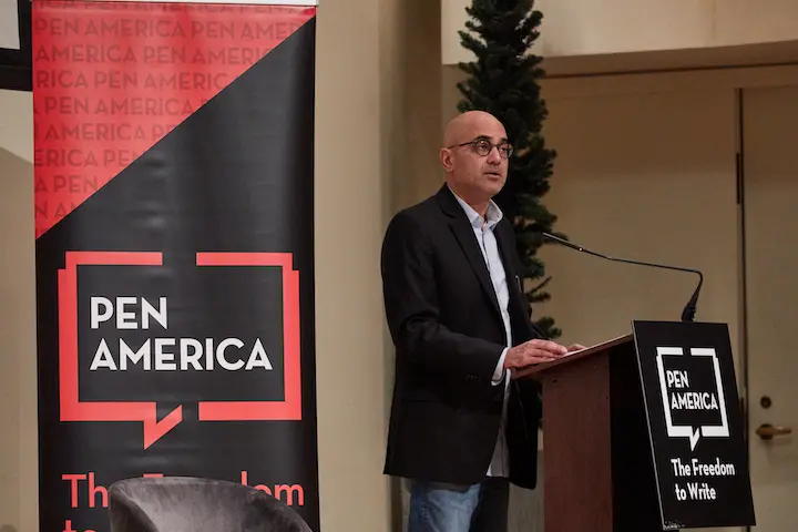 A person in a suit stands at a podium with an Ayad Akhtar PEN America Town Hall sign, speaking at an event. A black and red PEN America banner and a Christmas tree are visible in the background.