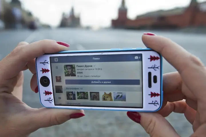 Close-up of hands with red nail polish holding a smartphone. The screen displays a VK profile, a popular social media platform in Russia, with the blurred outdoor background highlighting digital free expression.