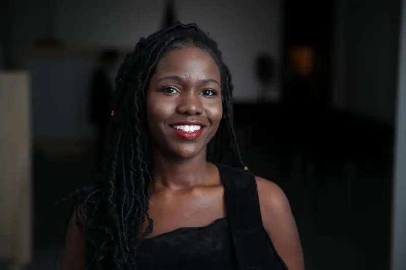 A woman with long, dark, braided hair smiles at the camera. She is wearing a sleeveless black top and stands in a dimly lit indoor setting, holding a copy of *The Path to Remembering* by M. Kiguwa.